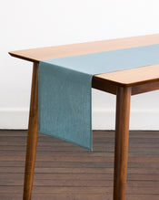 A wooden table with angled legs is topped with a Lagoon 100% French Flax Linen Table Runner by Bed Threads, draping over one end. Set on a wooden floor against a white wall, it brings simple elegance to your dining space. - view