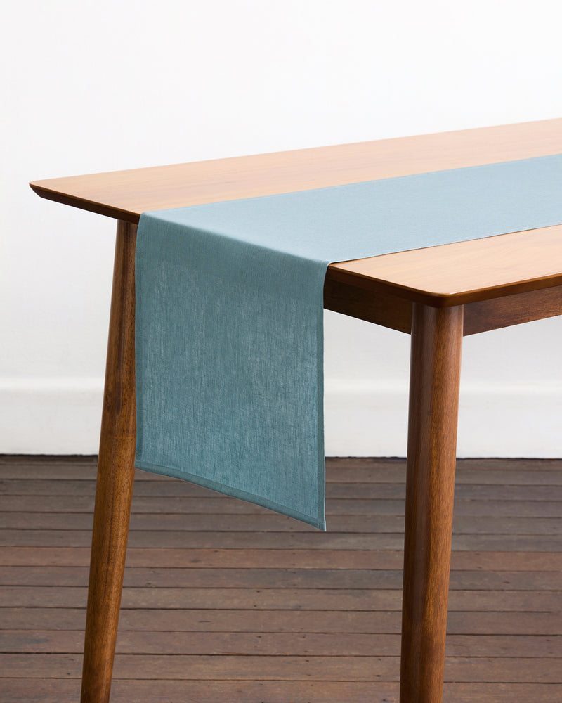 A wooden table with angled legs is topped with a Lagoon 100% French Flax Linen Table Runner by Bed Threads, draping over one end. Set on a wooden floor against a white wall, it brings simple elegance to your dining space.