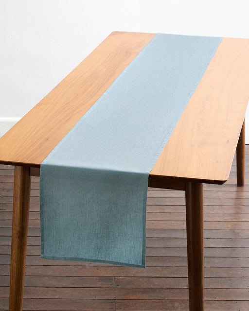 A Lagoon 100% French Flax Linen Table Runner by Bed Threads in light blue is neatly laid along the center of a rectangular wooden table with angled legs, set on a dark wood floor against a white wall.