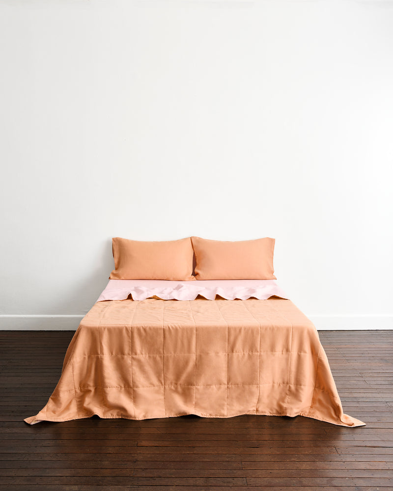 A neatly made bed with a double-sided peach-colored quilted coverlet in terracotta & rosewater, with two matching pillows in terracotta set against a plain white wall and dark wood floor.