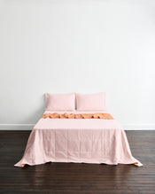 A neatly made bed with a double-sided light pink quilted coverlet in terracotta & rosewater, with two matching pillows in rosewater set against a plain white wall and dark wood floor. - view