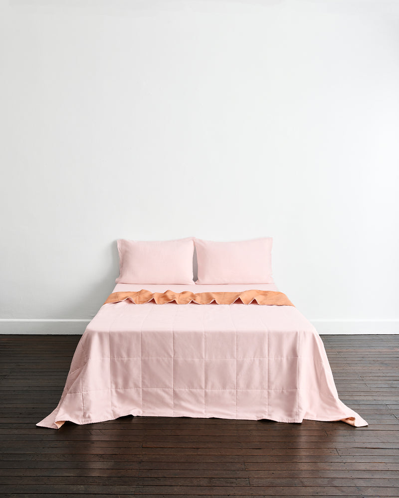 A neatly made bed with a double-sided light pink quilted coverlet in terracotta & rosewater, with two matching pillows in rosewater set against a plain white wall and dark wood floor.