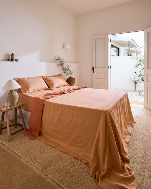 A cozy bedroom features the Bed Threads Terracotta & Hazelnut Two-Tone Quilt, a wooden side table with a lamp, a potted plant, and double doors to a sunny patio. Neutral tones and natural light create a calm, inviting feel.