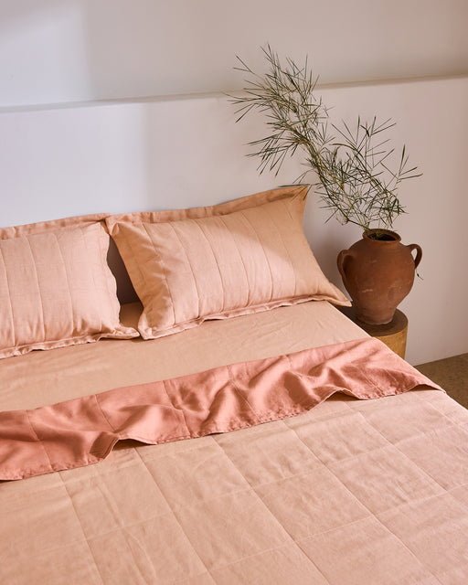 A neatly made bed with peach-colored bedding and Bed Threads Terracotta Quilted Pillowcases (Set of Two), crafted from soft French flax linen. A terracotta vase with green branches sits on a wooden stool by the white wall.