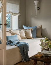 A cozy living room corner with a cream sofa, blue pillows, a Bed Threads Crème & Coast 100% French Flax Linen Swirl Cushion Cover, a light blue throw, wooden coffee table with flowers, and a window with sheer roman shade streaming in light. - view