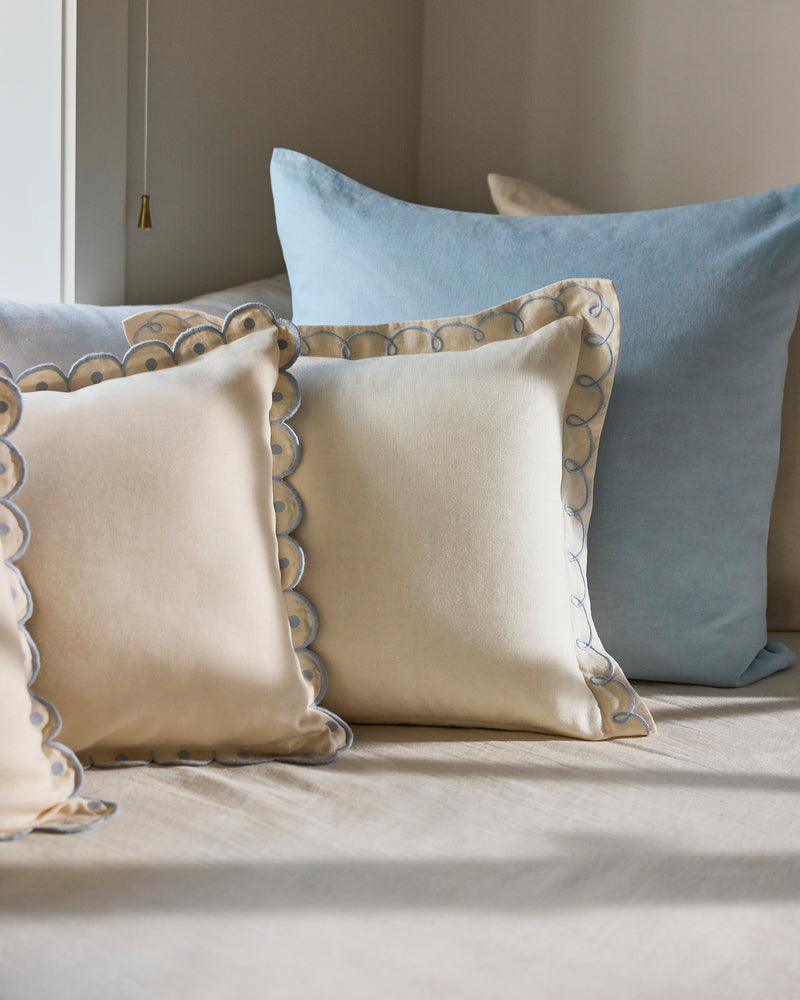 A close-up of a bed with two cream Bed Threads Crème & Coast 100% French Flax Linen Swirl Cushion Covers and one large light blue linen pillow, arranged against a beige wall.