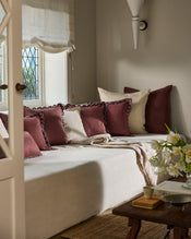 A cozy daybed with cream French flax linen bedding is styled with maroon and beige pillows near a patterned glass window. The inviting setting features the Bed Threads Cacao & Coast Swirl Cushion Cover, a soft throw, and a wooden table. - view