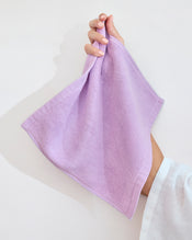 A hand displays a neatly folded Bed Threads Violet 100% Cotton Terry Face Washer (Set of Two), crafted from absorbent, skin-friendly fabric, against a plain white background. - view