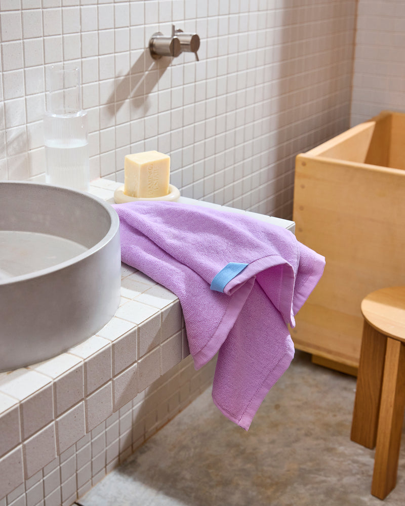 The Violet 100% Cotton Terry Hand Towel by Bed Threads sits on a white tiled sink beside a soap bar and glass of water, with a wooden stool and tub in the softly lit, quick-drying bathroom.