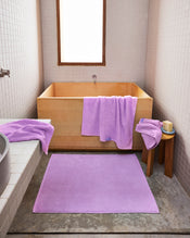 A modern bathroom features white tiled walls, a wooden soaking tub, and the Bed Threads Violet 100% Cotton Terry Bath Mat with light purple towels and washcloths. A stool beside the tub holds a towel, candle, and brush for added comfort. - view