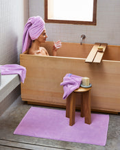 A person relaxes in a wooden bathtub, wearing a purple towel turban and holding a glass of water. Nearby, the Bed Threads Violet 100% Cotton Terry Bath Sheet is draped over a stool, with a matching bath mat on the floor. - view