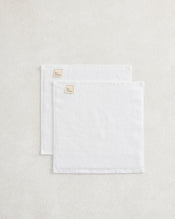 A set of two Crisp White 100% Cotton Terry Face Washers by Bed Threads, neatly folded with beige branded fabric labels in one corner, rests atop a light textured surface. - view