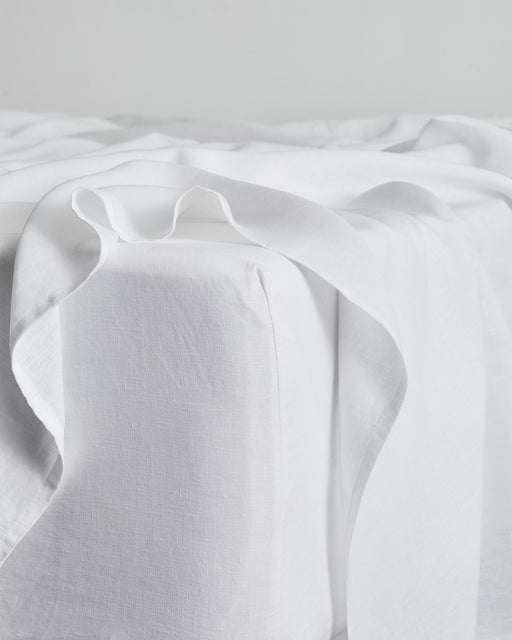 A close-up of the Bed Threads Coast, Lagoon & White 100% French Flax Linen Bedding Bundle shows soft, smooth white sheets draped over a mattress corner in a minimalistic setting, highlighting their luxurious texture.