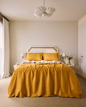 A cozy bedroom featuring Bed Threads’ Turmeric & Crème 100% French Flax Linen Swirl Pillowcases, a mustard linen duvet, white headboard and side tables with lamps, floral vase, white curtains, and a modern white ceiling light. - view