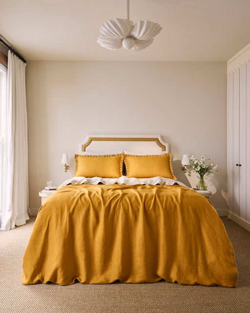 A cozy bedroom featuring Bed Threads’ Turmeric & Crème 100% French Flax Linen Swirl Pillowcases, a mustard linen duvet, white headboard and side tables with lamps, floral vase, white curtains, and a modern white ceiling light.