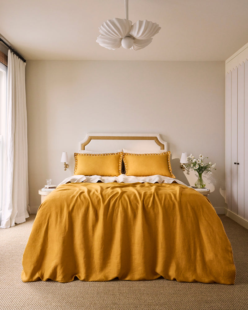A cozy bedroom featuring Bed Threads’ Turmeric & Crème 100% French Flax Linen Swirl Pillowcases, a mustard linen duvet, white headboard and side tables with lamps, floral vase, white curtains, and a modern white ceiling light.