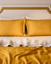 A neatly made bed with a beige and gold headboard, white sheets, a mustard yellow blanket, and Bed Threads Turmeric & Crème 100% French Flax Linen Swirl Pillowcases (Set of Two) in mustard yellow. - view
