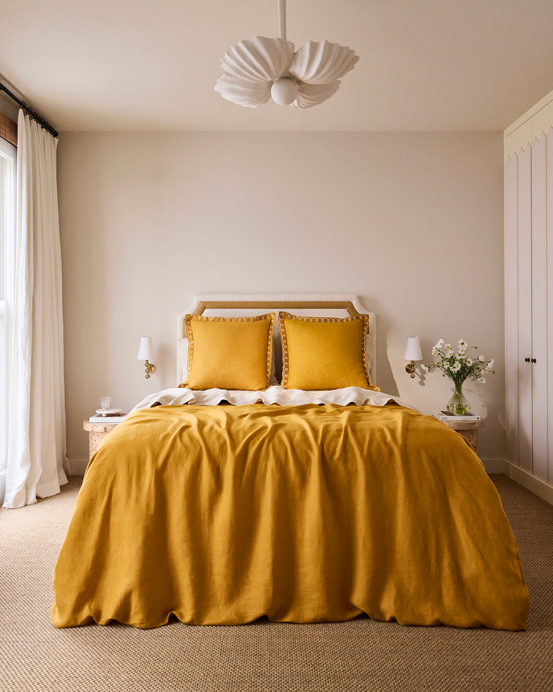 A neatly made bed features Bed Threads’ Turmeric & Crème 100% French Flax Linen Swirl European Pillowcases, paired with a mustard yellow comforter, against a beige wall. Two tables with lamps and flowers sit under a white pendant light.
