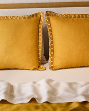 Two Bed Threads Turmeric & Crème 100% French Flax Linen Swirl European Pillowcases rest against a beige headboard. A white ruffled blanket is folded at the end, partly covering a 170GSM French flax linen mustard yellow bedspread. - view