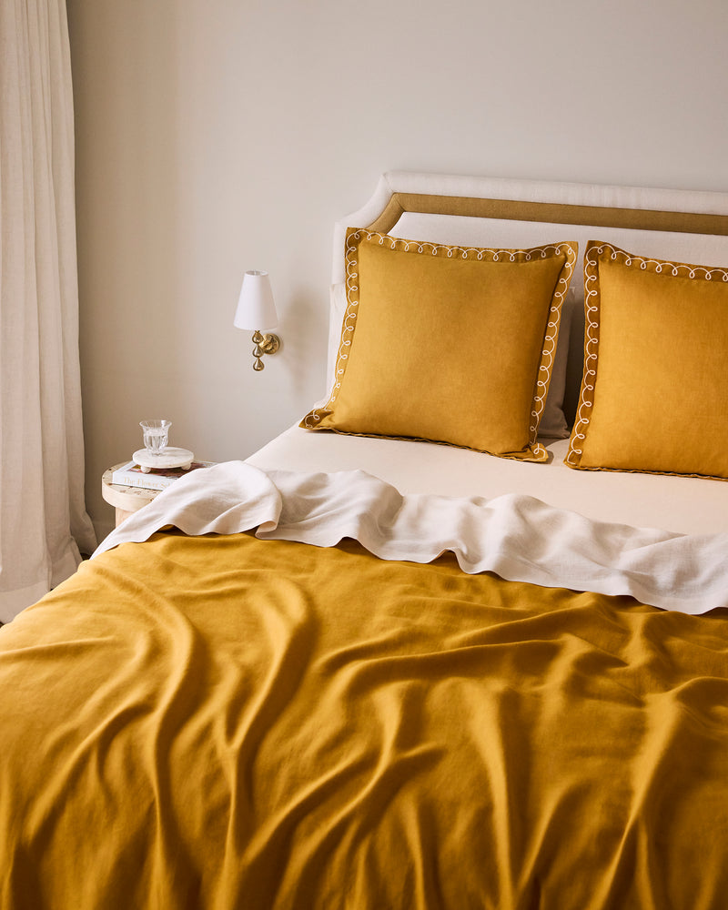 A neatly made bed features Bed Threads' Turmeric & Crème 100% French Flax Linen Swirl European Pillowcases, paired with a matching duvet and white throw. A round nightstand with a cup and saucer sits nearby, beneath a wall sconce.