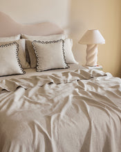 A neatly made bed features light beige French flax linen bedding with two Bed Threads Oatmeal & Charcoal 100% French Flax Linen Arch Cushion Covers, each with black scalloped edges, beside a sculptural white table lamp on the bedside table. - view