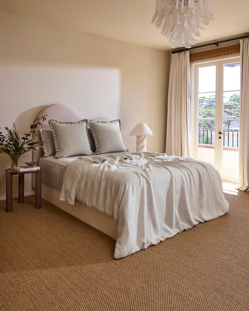 A cozy, sunlit bedroom with a neatly made bed featuring Bed Threads Oatmeal & Charcoal 100% French Flax Linen Swirl European Pillowcases, decorative pillows, a wooden nightstand, a lamp, and French doors with sheer curtains to a balcony.