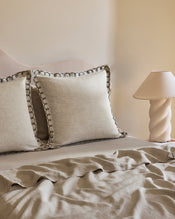 A neatly made bed with beige French flax linen and two Bed Threads Oatmeal & Charcoal 100% French Flax Linen Swirl European Pillowcases beside a small table topped with a cream spiral lamp and matching shade. - view