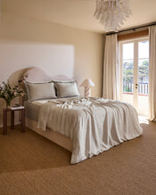 A cozy bedroom features a neatly made bed with Bed Threads’ Oatmeal & Charcoal 100% French Flax Linen Swirl Pillowcases, a small bedside table with a plant and lamp, and sheer curtains letting natural light in through a large window. - view