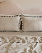 A neatly made bed with a beige upholstered headboard, two Bed Threads Oatmeal & Charcoal 100% French Flax Linen Swirl Pillowcases featuring black trim, and a matching slightly wrinkled French flax linen blanket. - view