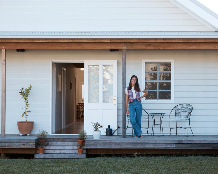 A woman stands and smiles on the porch of a light-colored house with potted plants, a black watering can, and a small outdoor table with two chairs. The front door is open, revealing part of the interior hallway.
