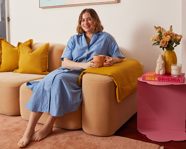 A woman in a blue dress sits barefoot on a beige sofa with yellow cushions, holding a mug. Beside her is a pink side table with books, a vase of yellow roses, and a lit candle in a cozy, colorful living room.