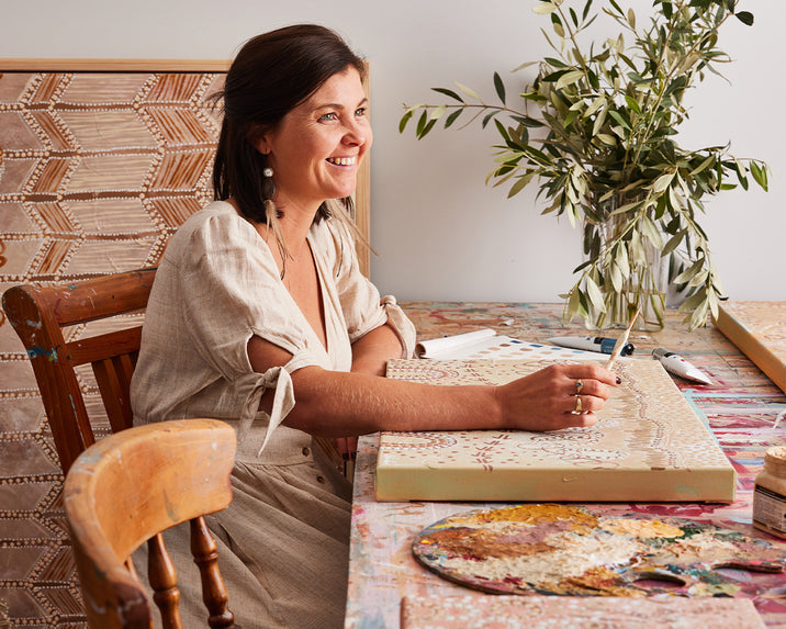 A woman in a light dress smiles while painting a patterned canvas at a colorful, paint-splattered table. Art supplies and a vase of leafy branches are nearby, and a finished artwork leans against the wall.