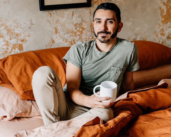 A man with short dark hair and a mustache sits on a bed with rust-colored bedding, wearing a green t-shirt and beige pants, holding a white mug and smiling slightly at the camera.
