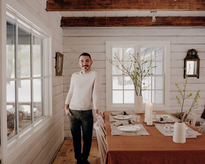 A person in a white sweater stands by a wooden dining table set for a meal in a cozy, rustic room with white shiplap walls, large windows, and simple decor, including candles and vases with branches.
