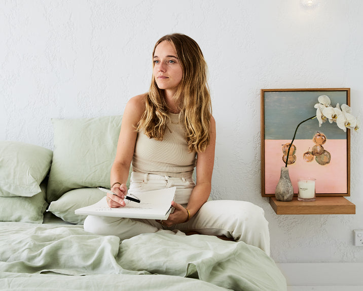 A woman with long hair, dressed in light-colored clothes, sits on a bed with pale green bedding, holding a notebook and pen. Beside her is a small shelf with a framed painting, a candle, and a white orchid.
