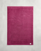 The Bed Threads Berry 100% Cotton Terry Bath Mat, a rectangular dark pink plush mat with a small white label on one corner, lies flat on a light gray surface. - view