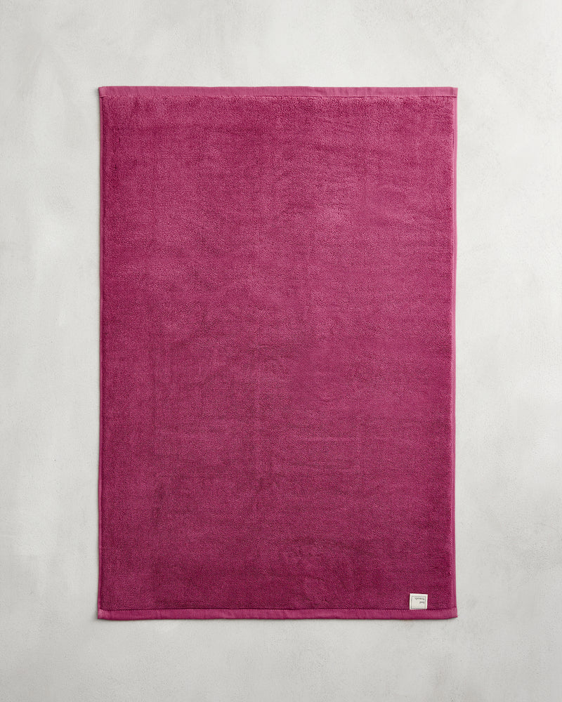 The Bed Threads Berry 100% Cotton Terry Bath Mat, a rectangular dark pink plush mat with a small white label on one corner, lies flat on a light gray surface.