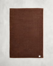 The Espresso 100% Cotton Terry Bath Mat by Bed Threads, a plain rectangular brown towel with a small white label in one corner, is laid flat on a light gray surface. - view