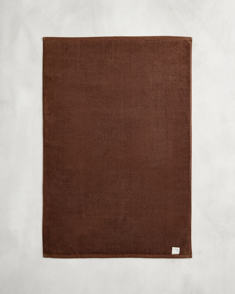 The Espresso 100% Cotton Terry Bath Mat by Bed Threads, a plain rectangular brown towel with a small white label in one corner, is laid flat on a light gray surface.