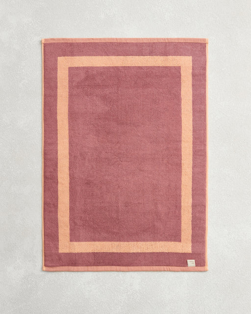 The Bed Threads Terracotta & Pink Clay Two-Toned 100% Cotton Terry Bath Mat, shown flat on a textured surface with a visible corner tag, pairs perfectly with plush towels for a coordinated bathroom style.