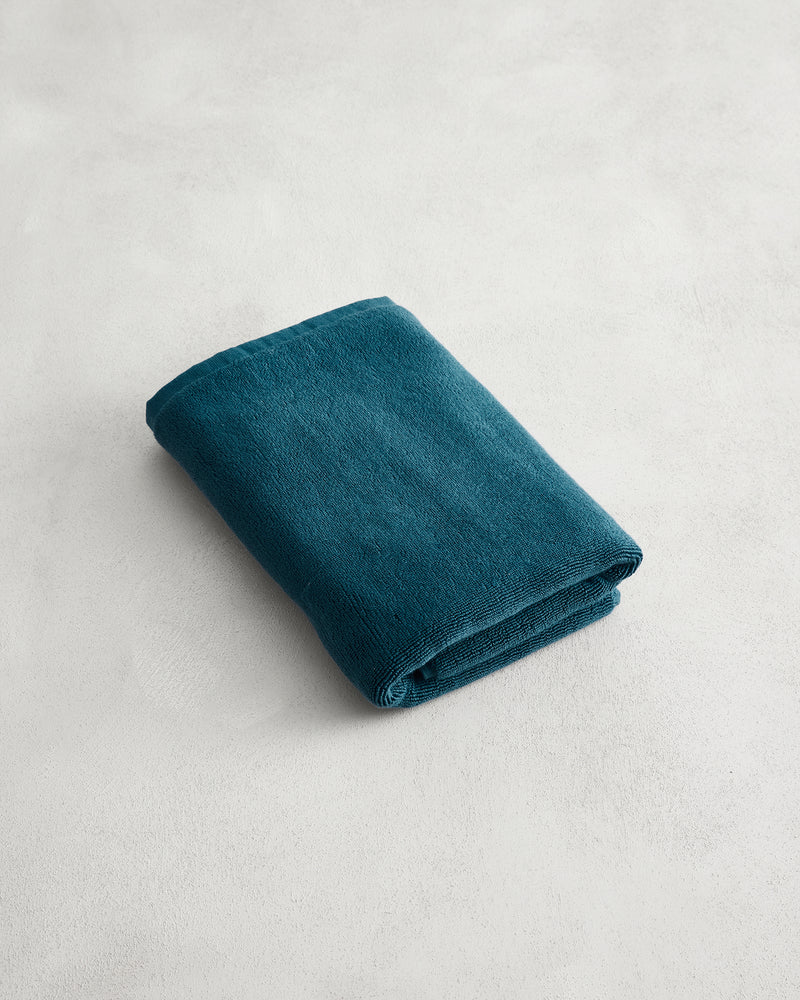 A neatly folded dark teal towel sits on a textured surface next to the Bed Threads Aegean 100% Cotton Terry Bath Mat, adding comfort and style.