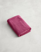 A plush Berry 100% Cotton Terry Bath Mat by Bed Threads lies on a light gray, textured surface next to a neatly folded, soft magenta towel. - view