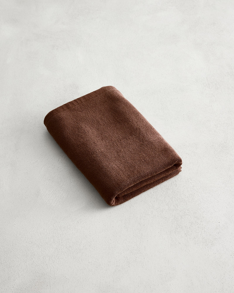 A neatly folded brown towel rests on a light gray, subtly textured surface next to the Bed Threads Espresso 100% Cotton Terry Bath Mat.