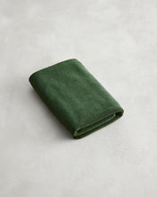 A neatly folded Moss 100% Cotton Terry Bath Mat by Bed Threads in dark green rests on a light gray textured surface, adding comfort and style. - view