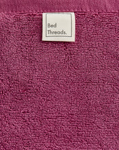A close-up of the Berry 100% Cotton Terry Bath Mat by Bed Threads shows its textured magenta surface and a sewn-on white label reading "Bed Threads." in black text, highlighting its plush feel and absorbency. - view