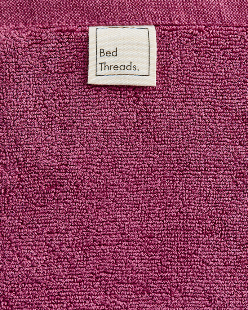 A close-up of the Berry 100% Cotton Terry Bath Mat by Bed Threads shows its textured magenta surface and a sewn-on white label reading 