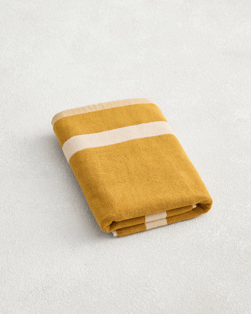 The Bed Threads Crème & Turmeric Two-Toned 100% Cotton Terry Bath Mat features mustard yellow and white stripes, adding a cozy accent to your bathroom when paired with plush towels on a light, textured surface.