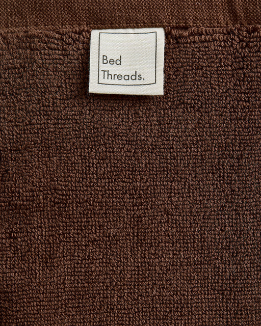 A close-up of the Espresso 100% Cotton Terry Bath Mat by Bed Threads, featuring a brown textured surface and a white label at the top with Bed Threads in black text.
