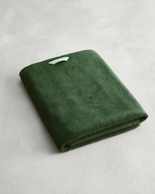 The Moss 100% Cotton Terry Bath Sheet by Bed Threads, in deep green with a white tag, rests on a light gray surface—ideal for anyone seeking plush, quick-drying towels.