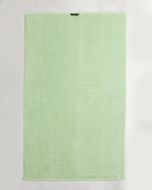 A set of two Apple 100% Cotton Terry Bath Sheets by Bed Threads, in light green, each rectangular with a dark green loop near the top edge and a white tag at the bottom right, laid flat on a white surface.